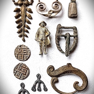 Könnte beinhalten: Eine Sammlung antiker Metallanhänger, Münzen und Schnallen in verschiedenen Formen und Größen. Zu den Artikeln gehören ein blattförmiger Anhänger, eine Männerfigur und mehrere runde Münzen mit geprägten Designs. Die Farben sind hauptsächlich Bronze und Silber.