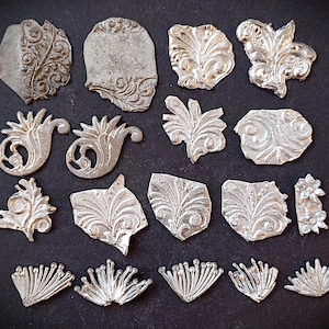 Könnte beinhalten: Eine Sammlung von siebzehn silberfarbenen Metallverzierungen mit verschiedenen geprägten Blumen-, Blatt- und Fächermustern. Diese kleinen, flachen Dekorationselemente weisen komplizierte erhabene Details und unregelmäßige Formen in vier Reihen auf.