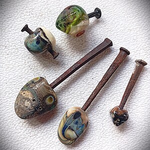 May include: Artisan-crafted glass beads with nail stems. The beads feature a variety of colors and patterns, including swirls of blue, green, and brown. The nail stems are made of rusted metal. The beads vary in size and shape.