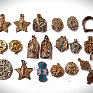 May include: A collection of twenty assorted metal charms and pendants in various shapes including stars, circles, and rectangles. The items feature embossed figures, religious imagery, and one blue enamel piece, all showing a weathered, aged patina.