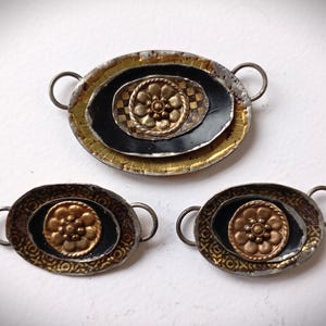 Puede incluir: Tres componentes de joyería metálicos de forma ovalada. El más grande tiene un diseño dorado y negro en capas con una flor dorada central. Los componentes más pequeños tienen diseños similares. Cada pieza tiene dos pequeños bucles para la fijación.
