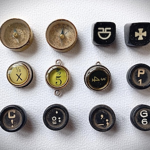 May include: A collection of twelve small objects including two brass compasses, two black square buttons with white symbols, and eight circular typewriter keys. The keys feature various characters including 1/4 X, % 5, 4 $, P 1, C !, a colon, a semicolon, and G 6, all arranged in a grid pattern.