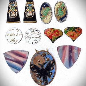 Könnte beinhalten: Eine Sammlung von Schmuckkomponenten aus Metall im Vintage-Stil. Enthält schwarz-goldene Anhänger, ovale und tropfenförmige Teile mit floralen und gemusterten Designs sowie einen Anhänger mit Schmetterling und Sternen. Die Farbpalette umfasst Schwarz, Gold, Blau, Rot und Weiß.