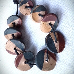 May include: A necklace of ten round, hand-painted ceramic beads. Each bead is divided into three sections of color: tan, dark green, and maroon. The beads are strung together with black cord, creating a unique, earthy accessory.