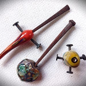 May include: Three handcrafted glass beads with metal accents. One is a vibrant red teardrop shape, another is a speckled brown and green orb, and the third is a yellow and blue orb with a central black hole. Each bead has a rusty brown metal rod.