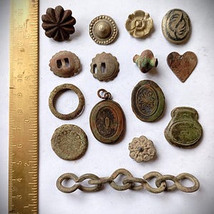Könnte beinhalten: Eine Sammlung verschiedener Metallobjekte, darunter Knöpfe, Anhänger und ein Kettenglied, angeordnet neben einem Messinglineal mit Zoll- und Zentimeterangaben. Die Gegenstände weisen verwitterte Texturen sowie kreisförmige, florale und herzförmige Formen auf.