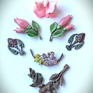 Könnte beinhalten: Eine Sammlung von Vintage-Blumenbroschen und -Anhängern in verschiedenen Designs. Die Stücke zeigen rosa, lila und braune Blumen mit grünen Blättern. Einige haben Perlenakzente. Der Schmuck ist auf einer weißen Oberfläche angeordnet.