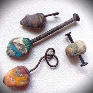 May include: Three handcrafted, rustic-style beads with metal accents. The beads are multi-colored, featuring shades of blue, orange, yellow, and brown. One bead is pierced by a nail, while the others have wire loops.