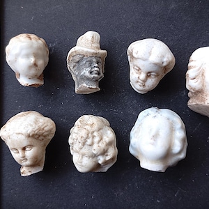 Puede incluir: Colección de siete cabezas de muñecas antiguas de porcelana, cada una con rasgos faciales y peinados únicos. Las cabezas son principalmente blancas con tonos envejecidos, y están sobre una superficie oscura. Una regla está en el fondo.