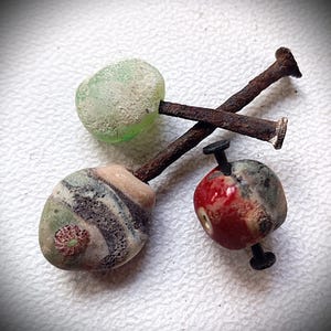 May include: Three antique glass beads in green, red, and patterned designs are pierced by rusty nails. The beads have a weathered appearance, suggesting age. The composition is set against a white background.