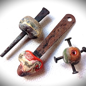 May include: Three handcrafted rustic objects. One features a nail with a brown and cream-colored bead. Another has a rusty metal strip with a red, white, and black swirled bead. The third has a bead with four nails.