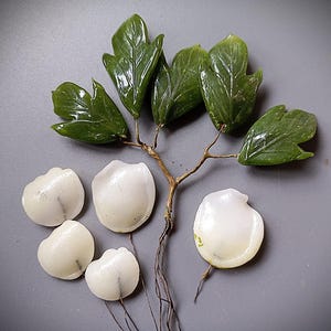 Könnte beinhalten: Ein dekorativer Zweig mit fünf glänzenden, dunkelgrünen, geschnitzten Steinblättern an dünnen braunen Drahtstielen, ergänzt durch vier glatte, abgerundete, muschelartige weiße Teile, die ebenfalls an dünnen braunen Drahtstielen befestigt sind.