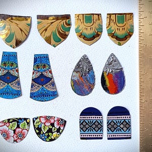 Könnte beinhalten: Eine Sammlung farbenfroher, dekorativer Ohrringkomponenten in verschiedenen Formen. Die Komponenten umfassen Schild-, Tropfen- und Bogenformen mit floralen, geometrischen und abstrakten Designs. Farben sind Blau, Gold, Rosa und Schwarz. Ein Lineal ist im Hintergrund.