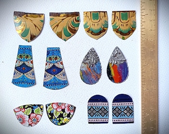 Puede incluir: Una colección de componentes de pendientes decorativos y coloridos en varias formas. Los componentes incluyen formas de escudo, lágrima y arco, con diseños florales, geométricos y abstractos. Los colores incluyen azul, dorado, rosa y negro. Una regla está en el fondo.