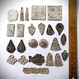 Puede incluir: Una colección de varios dijes y colgantes de metal plateado con patrones en relieve, incluyendo espirales, animales y motivos florales, dispuestos junto a una regla de metal que muestra medidas de hasta 15 cm como referencia.