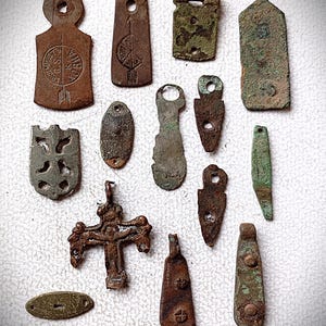 Könnte beinhalten: Eine Sammlung antiker Metallanhänger und -charms in verschiedenen Formen und Größen. Die Stücke sind hauptsächlich in Braun- und Grüntönen gehalten und weisen deutliche Alters- und Gebrauchsspuren auf. Einige zeigen dekorative Designs und Symbole.