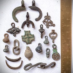 Peut inclure: Une collection de pendentifs et de breloques antiques en bronze, comprenant diverses formes telles que des croissants de lune, des gouttes et des motifs ornés. Les pièces montrent des signes d'âge, avec une règle en centimètres pour l'échelle.