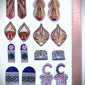 Könnte beinhalten: Eine Sammlung von kunstvollen, antiken Schmuckstücken in verschiedenen Formen und Größen. Die Stücke weisen rote, goldene, blaue und weiße Emaille-Details auf. Einige haben florale und geometrische Muster. Ein Lineal ist zum Maßstab in Zentimetern enthalten.