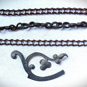 Puede incluir: Un surtido de componentes de ferretería metálicos antiguos, que incluyen tres cadenas de diferentes diseños y un soporte curvo decorativo con detalles florales. Las cadenas son de color marrón óxido, y el soporte es de metal más oscuro y envejecido.