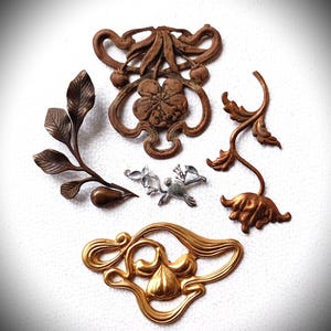 Könnte beinhalten: Eine Auswahl antiker Metallverzierungen in verschiedenen floralen und botanischen Designs. Die Teile umfassen eine bronzefarbene Blume, ein goldfarbenes Blatt und einen silberfarbenen Vogel. Diese dekorativen Elemente eignen sich ideal für Bastelarbeiten und Schmuckherstellung.