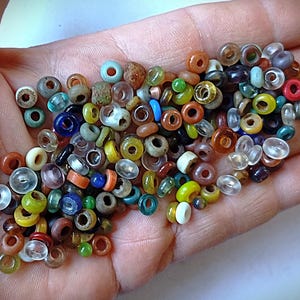 May include: A collection of small, round, multi-coloured glass beads in various shades including blue, yellow, green, red, clear, and brown. These beads feature a central hole and exhibit diverse textures and finishes, ranging from translucent to opaque, scattered together in a pile.