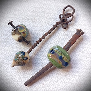 May include: Artisan-crafted glass beads with intricate designs in shades of green, blue, and brown, mounted on repurposed nails and twisted metal. The beads have a unique, organic shape, and the metal elements show a rustic, aged appearance.