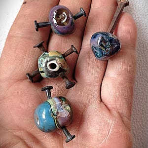 May include: Four handmade glass beads with nail accents. The beads are various colors, including blue, purple, green, and yellow, with unique patterns and textures. The nails are dark metal, creating a striking contrast with the colorful glass.
