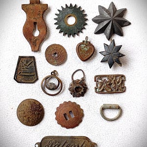 Könnte beinhalten: Eine Sammlung antiker Metallobjekte, darunter eine Schlüssellochform, Sternausbrüche, ein Herz und ein Ring. Die Objekte sind in verschiedenen Braun- und Bronzetönen gehalten und weisen Alters- und Abnutzungserscheinungen auf. Ein rechteckiges Objekt mit dem Wort "Patent" ist ebenfalls enthalten.