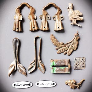 Könnte beinhalten: Eine Auswahl antiker Metallbeschläge in verschiedenen Formen und Größen. Enthält dekorative Griffe, blattförmige Griffe, eine Vogelfigur, ein federartiges Ornament sowie kleine rechteckige und quadratische Teile. Zwei ovale Etiketten lauten "hier rein" und "da raus".