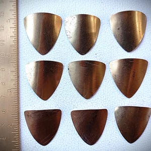 Könnte beinhalten: Neun bronzefarbene Gitarrenplektren, angeordnet in einem Raster. Jedes Plektrum hat eine abgerundete Oberseite und eine spitze Unterseite. Ein Lineal mit Zentimetermarkierungen ist links im Bild zu sehen.
