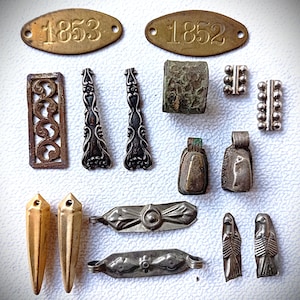 Könnte beinhalten: Eine Sammlung antiker Schmuckkomponenten und -funde aus Metall. Enthält Messinganhänger mit den Zahlen 1853 und 1852, verzierte Filigranstücke, Anhänger und dekorative Elemente in Silber- und Goldtönen.