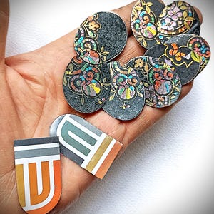 May include: A selection of decorative metal embellishments with detailed floral and geometric patterns. The pieces have a dark grey base with colourful accents of red, yellow, blue, and green. Some are oval, others are rectangular.