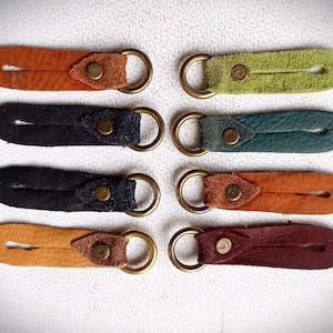 Può includere: Una collezione di sette portachiavi in pelle in vari colori, tra cui arancione, verde, nero e bordeaux. Ogni portachiavi presenta un anello in ottone e un dettaglio rivettato. Disposti su una superficie bianca.