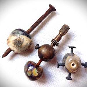 May include: Three unique, handcrafted objects. One features a rusty nail through a marbled, cream-colored bead with black and gray swirls. Another has a brass ball with a gear-like component. The third has a bead with four small, dark metal posts.