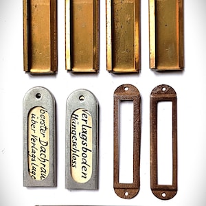 Puede incluir: Un conjunto de diez piezas de herrajes metálicos que incluye cuatro portaetiquetas rectangulares de latón con orificios, dos portaetiquetas plateados con etiquetas "Verlagsboden Hängeschloss" y "berster Dachraum über Verlagslage", dos marcos rectangulares de color marrón oscuro y dos tiras largas de latón.