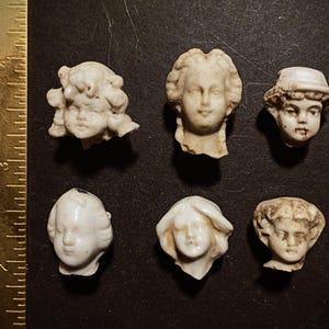 Puede incluir: Seis cabezas de muñecas antiguas de porcelana, en varios estilos, dispuestas sobre una superficie oscura. Las cabezas son de color blanquecino, con rasgos faciales y peinados detallados. Una regla es visible en el lado izquierdo.