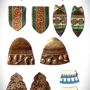 May include: A collection of assorted metal and ceramic jewelry components including two floral-shaped pieces, two shield-shaped pieces with red and green patterns, two pointed pieces, two brass-toned semi-circles, two textured shapes, and two blue and white curved segments.