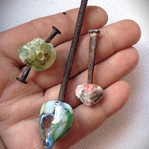 May include: Three handmade nail pendants with unique glass beads. One is a light green, another is a mix of blue, green, and white, and the third is red and white. The nails are rusty, adding a rustic charm to the jewelry.