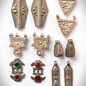 Op de afbeelding: Een verzameling antieke zilveren hangers en bedels. De collectie bevat ruitvormige, driehoekige en rechthoekige hangers, sommige met decoratieve patronen en gekleurde stenen accenten. De bedels zijn gerangschikt op een witte ondergrond.