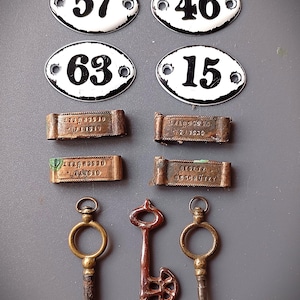 Könnte beinhalten: Sammlung von Vintage-Metallgegenständen, darunter vier ovale, weiße Adressschilder mit schwarzen Zahlen, vier kleine rechteckige Metallanhänger und drei antike Schlüssel. Die Schilder zeigen die Zahlen 57, 46, 63 und 15.