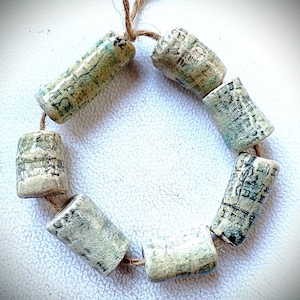 May include: A necklace of cylindrical beads with a crackle glaze in shades of green, blue, and cream. The beads are strung on a natural twine, creating a circular shape. The beads have a textured surface and a rustic appearance.