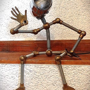 Könnte beinhalten: Einzigartige Metallskulptur einer Figur, die auf einem Holzregal sitzt. Der Kopf ist ein Chromscheinwerfer mit Messinggelenken und -händen. Der Körper besteht aus Metallrohren und Federn, was ein industrielles Kunstwerk ergibt.