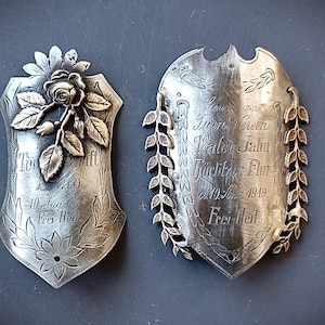 Puede incluir: Dos placas de plata antiguas con textos grabados y diseños florales. Una placa presenta un motivo de rosa y hojas, mientras que la otra tiene un borde de laurel. Ambas placas tienen texto y fechas en alemán.