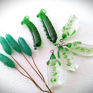 Puede incluir: Una colección de dijes de vidrio que incluye cuatro piezas texturizadas en forma de pepino verde sobre alambres de cobre, dos colgantes de pepino verde oscuro y tres vainas de guisantes de vidrio transparente con guisantes verdes.