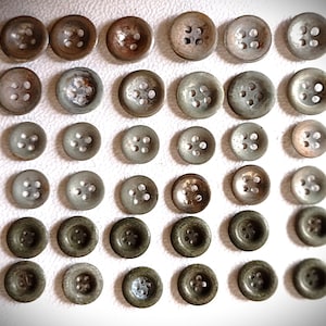 Könnte beinhalten: Eine Sammlung von Vintage-Metallknöpfen in verschiedenen Größen und Grautönen. Jeder Knopf hat vier Löcher zum Annähen. Die Knöpfe sind in ordentlichen Reihen angeordnet und zeigen ihre unterschiedlichen Texturen und Oberflächen.