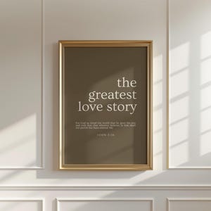 May include: A framed print with the text "the greatest love story" in white on a brown background. Below the title is a Bible verse from John 3:16. The frame is gold.
