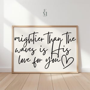 May include: A framed print with the words "mightier than the waves is His love for you" in black script. The frame is light wood, and the background is off-white. A heart symbol is at the end of the text.