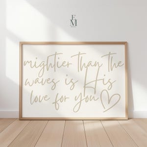 May include: A framed print with the text "Mightier Than The Waves Is His Love For You" in a light brown script font. The print has a cream-colored background and a wooden frame. The print is displayed on a light wood floor.