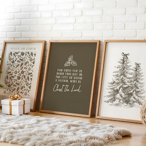 May include: Three framed art prints with wooden frames lean against a white brick wall. The prints feature botanical patterns, text, and Christmas tree illustrations. A white gift box with a gold bow sits in front of the prints.