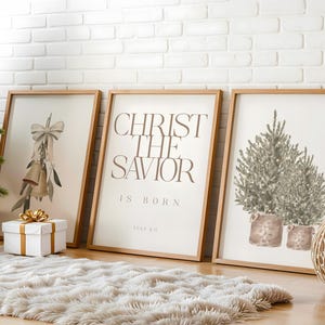 May include: Three framed art prints with wooden frames lean against a white brick wall. The prints feature watercolour illustrations of Christmas bells, text that reads "Christ the Saviour is Born", and potted Christmas trees. A white gift box with a gold bow sits in front.